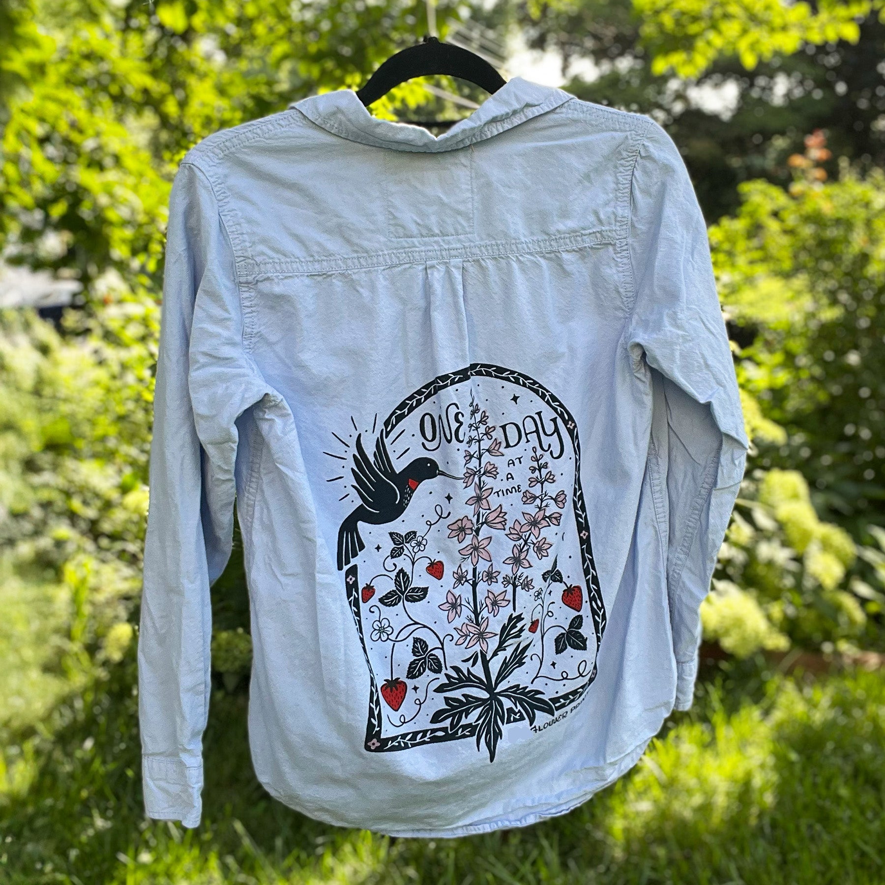 Light blue denim jacket with a graphic design on a hanger against a green outdoor background. July birth flower and fruit, larkspur & strawberries with a hummingbird. Hand-pulled screenprinted thrifted apparel with original Cornflower Print Co. illustration.
