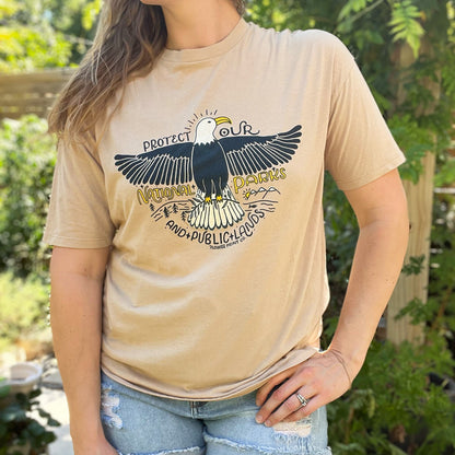 Person wearing a beige t-shirt with an eagle graphic and text outdoors. Protect Our National Parks. hand-pulled screenprinted illustration on thrifted apparel, handmade, upcycled apparel