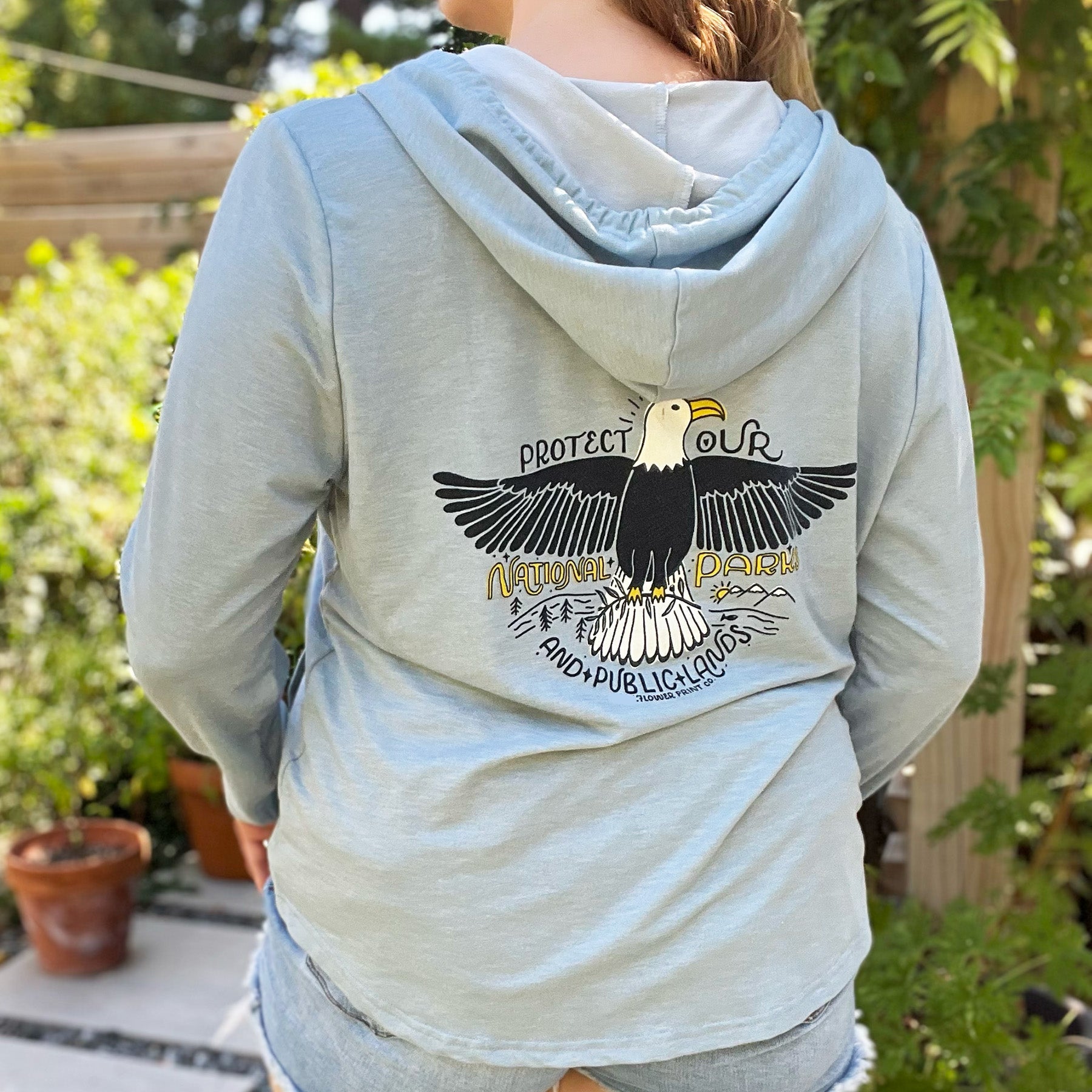 Person wearing a light gray hoodie with an eagle design on the back, standing outdoors. Protect Our National Parks. hand-pulled screenprinted illustration on thrifted apparel, handmade, upcycled apparel