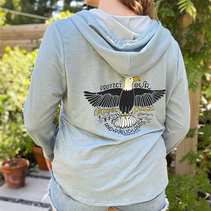 Person wearing a light gray hoodie with an eagle design on the back, standing outdoors. Protect Our National Parks. hand-pulled screenprinted illustration on thrifted apparel, handmade, upcycled apparel