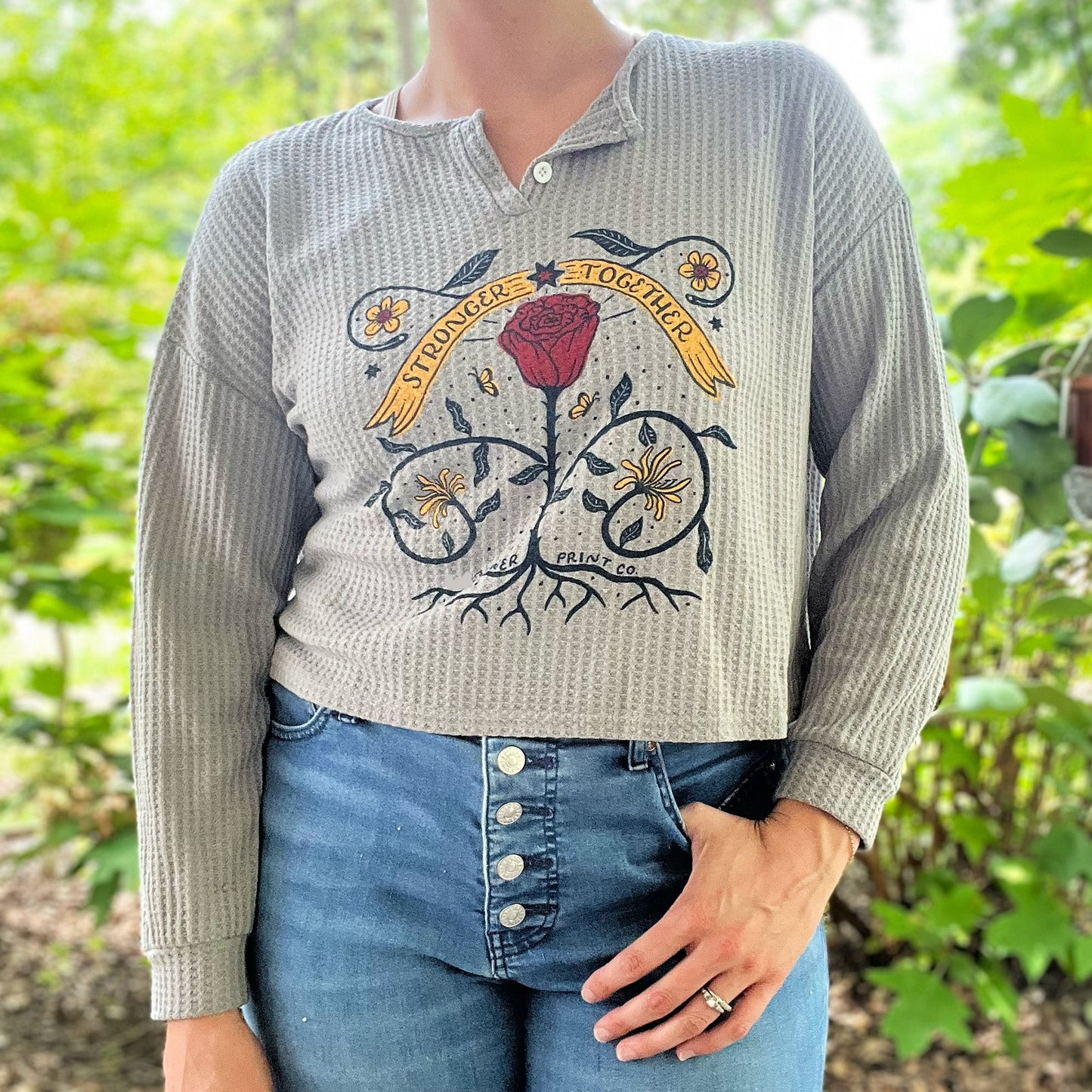 Person wearing a waffle knit cropped long sleeve shirt with a floral design and text in a garden setting. June birth flower of rose and honeysuckle with the phrase Stronger Together message. Hand-pulled screenprinted thrifted apparel with original illustration by Cornflower Print Co.