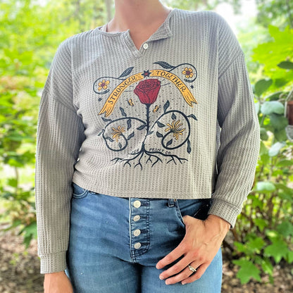Person wearing a waffle knit cropped long sleeve shirt with a floral design and text in a garden setting. June birth flower of rose and honeysuckle with the phrase Stronger Together message. Hand-pulled screenprinted thrifted apparel with original illustration by Cornflower Print Co.