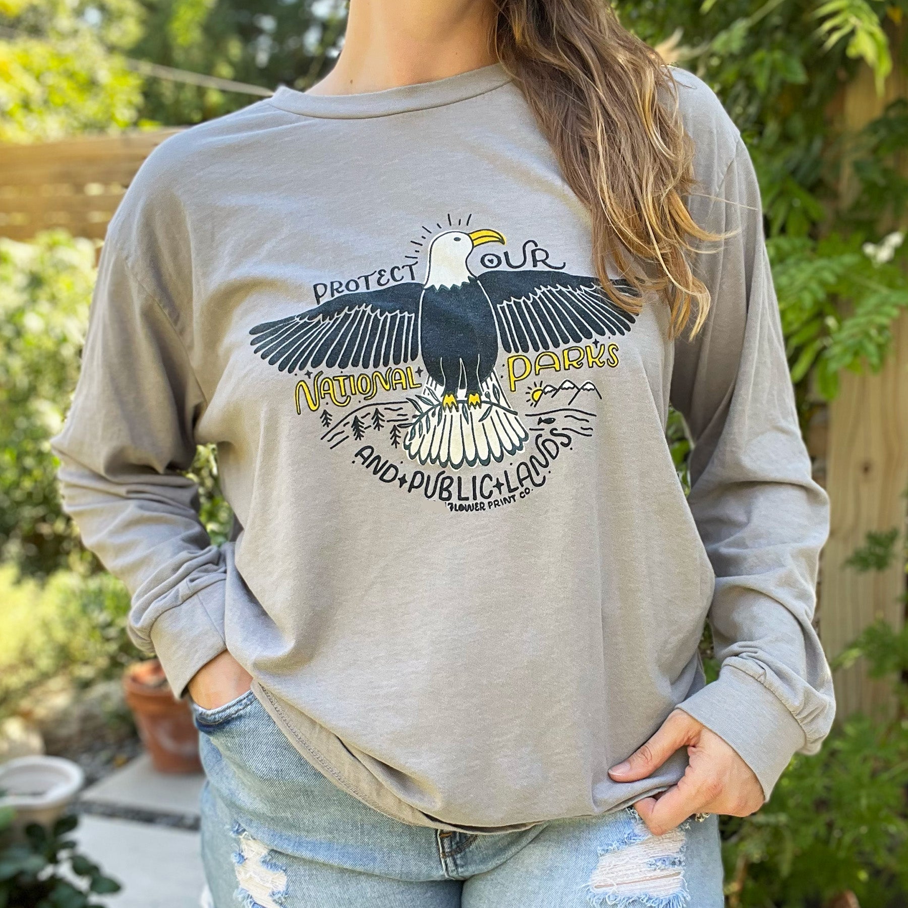 Person wearing a gray long sleeve shirt with an eagle graphic and text in a garden setting. Protect Our National Parks. hand-pulled screenprinted illustration on thrifted apparel, handmade, upcycled apparel