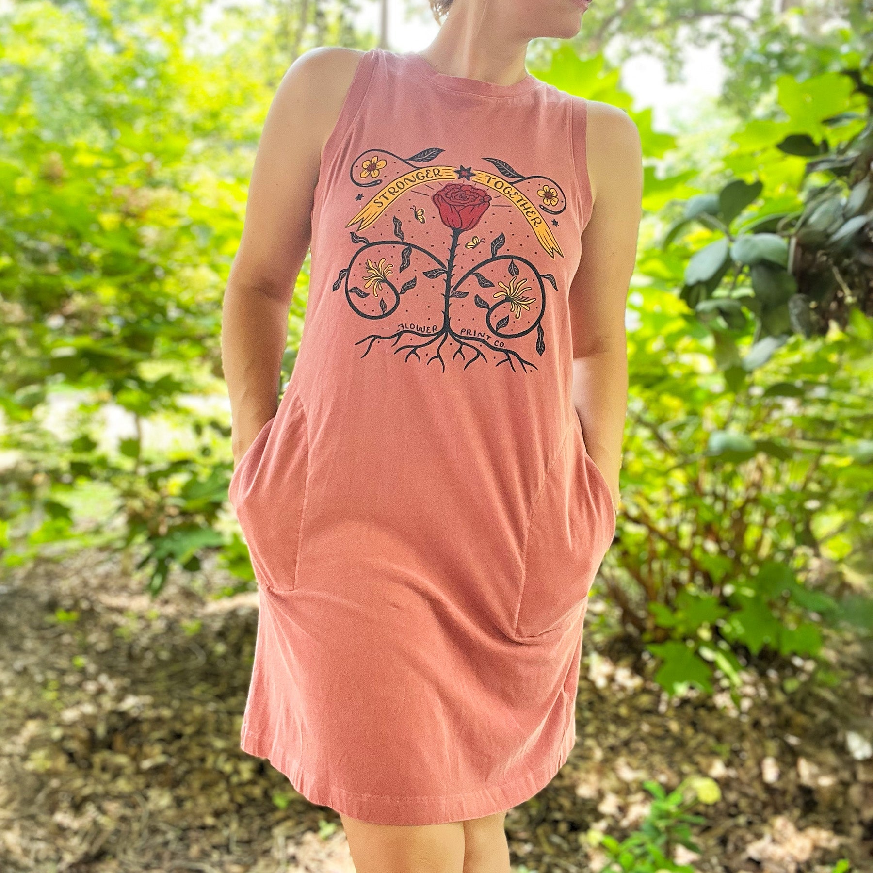 Person wearing a pink sleeveless dress with pockets with a graphic design in a forest setting. June birth flower of rose and honeysuckle with the phrase Stronger Together message. Hand-pulled screenprinted thrifted apparel with original illustration by Cornflower Print Co.
