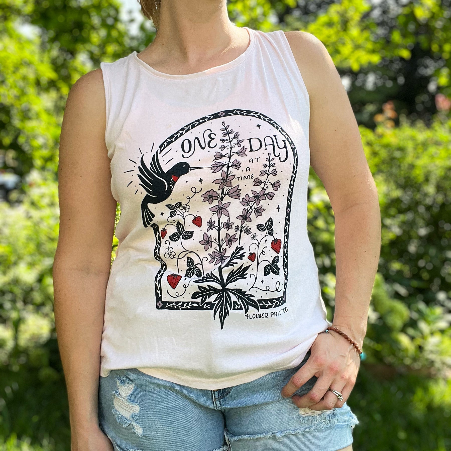 Person wearing a light pink tank top with a graphic design outdoors. July birth flower and fruit, larkspur & strawberries with a hummingbird. Hand-pulled screenprinted thrifted apparel with original Cornflower Print Co. illustration.