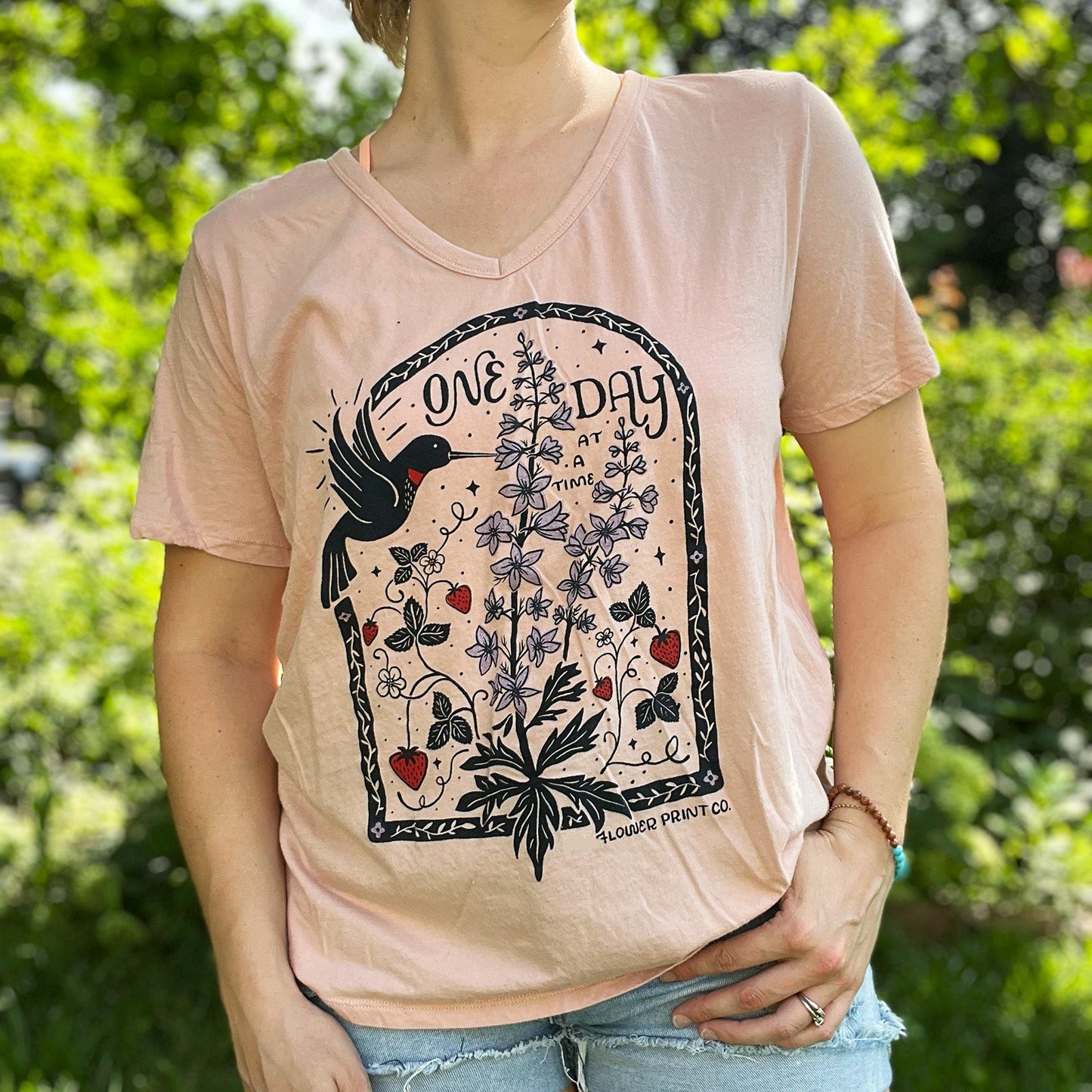 Person wearing a pink t-shirt with nature-themed design outdoors. July birth flower and fruit, larkspur & strawberries with a hummingbird. Hand-pulled screenprinted thrifted apparel with original Cornflower Print Co. illustration.