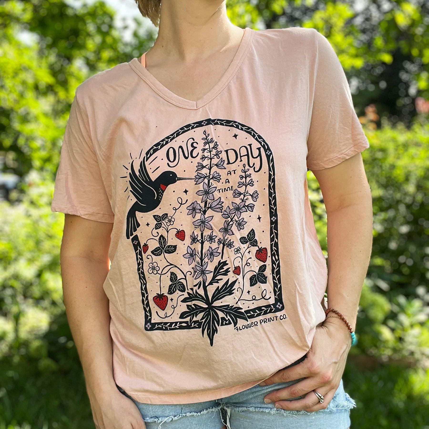 Person wearing a pink t-shirt with nature-themed design outdoors. July birth flower and fruit, larkspur & strawberries with a hummingbird. Hand-pulled screenprinted thrifted apparel with original Cornflower Print Co. illustration.