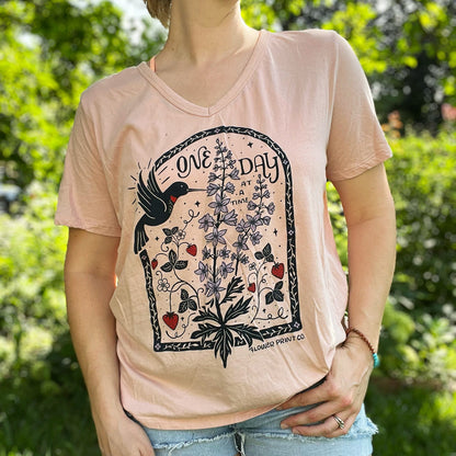 Person wearing a pink t-shirt with nature-themed design outdoors. July birth flower and fruit, larkspur & strawberries with a hummingbird. Hand-pulled screenprinted thrifted apparel with original Cornflower Print Co. illustration.