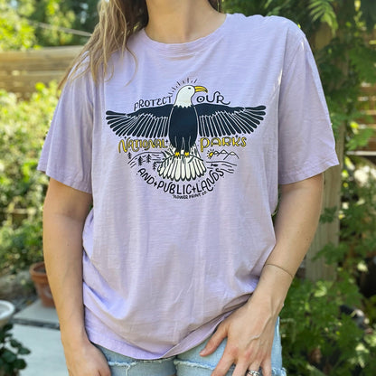 Person wearing a light purple t-shirt with an eagle graphic and text outdoors. Protect Our National Parks. hand-pulled screenprinted illustration on thrifted apparel, handmade, upcycled apparel