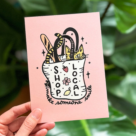 A postcard with a pink background, featuring a cartoon illustration of a cat inside a tote bag, alongside the text 'Shop Local' and some floral elements. The postcard is held in a hand with a green leafy background.