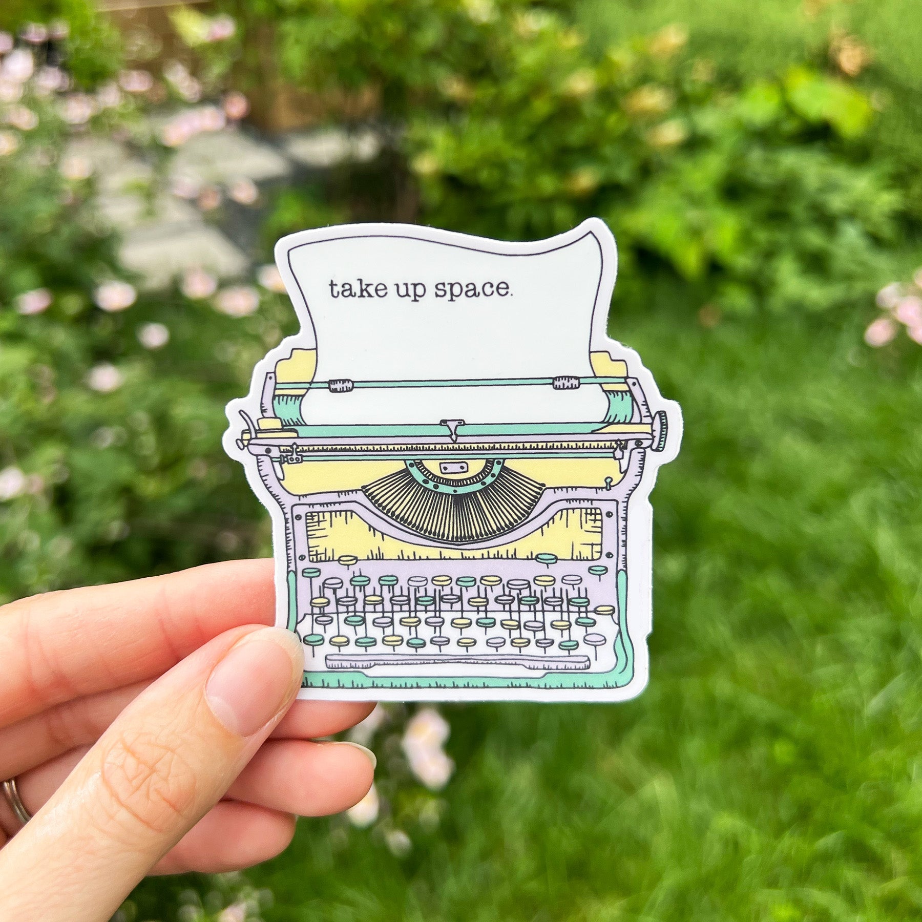 A vinyl sticker with a typewriter illustration and the phrase 'Take Up Space' written on it, held against an outdoor background.